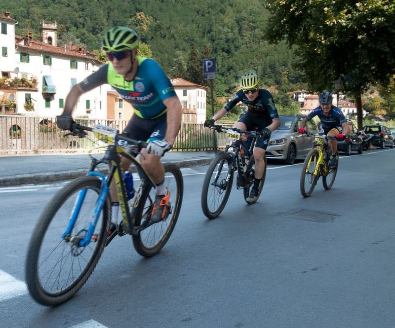 Three mountain-bike racers rounding a bend on the road beside the river
